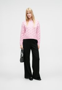 Suéter de punto rosa claro con patrón texturizado, combinado con pantalones negros de pierna ancha. La modelo sostiene un pequeño bolso negro. Fondo neutral.