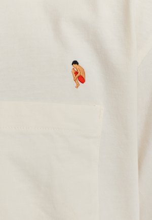 Crème katoen t-shirt met een kleine, kleurrijke geborduurde figuur in een hurkende positie, met een rode accent en zwart haar. Inclusief een detail van een zak.