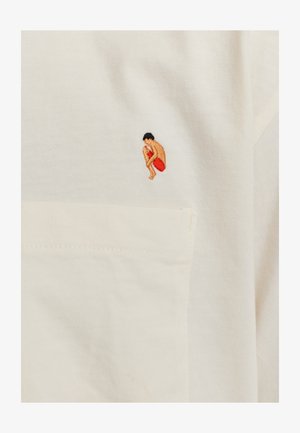 Crème katoen t-shirt met een kleine, kleurrijke geborduurde figuur in een hurkende positie, met een rode accent en zwart haar. Inclusief een detail van een zak.