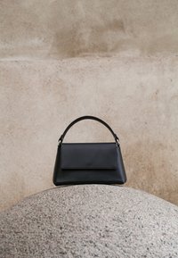 Μαύρο δερμάτινο τσαντάκι με κορυφαία λαβή τοποθετημένο πάνω σε μια υφή στρογγυλής πέτρας, με φόντο έναν μπεζ τσιμεντένιο τοίχο.