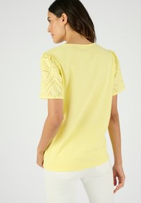 Geel t-shirt met korte pofmouwen en oogjesdetails. Zachte katoenen stof met een losse pasvorm, ronde halslijn en een gladde textuur.