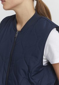Mörkblå quiltad väst med ribbad krage, framdragkedja och öppna ärmhål, para med en vit T-shirt under. Slät, lätt textur.