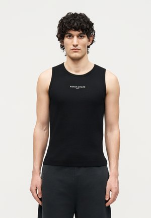 Homme aux cheveux bouclés portant un débardeur noir avec le texte « MAISON KITSUNÉ PARIS », debout devant un fond clair uni.