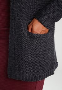 Mano en el bolsillo de un cárdigan de punto texturizado gris oscuro sobre un fondo de tela ribeteada burdeos.