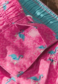 Rožiniai �šortai su mėlynais flamingo raštais, elastiniu juosmeniu ir maža kišene, uždaryta Velcro užtrauktuku.