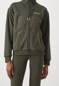 Giacca in pile con zip di colore verde scuro, con tessuto liscio a contrasto sulle spalle. Presenta dettagli gialli, tasche frontali e polsini a coste.