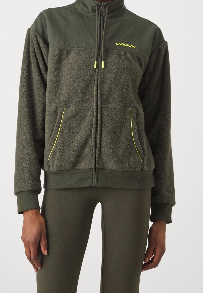 Giacca in pile con zip di colore verde scuro, con tessuto liscio a contrasto sulle spalle. Presenta dettagli gialli, tasche frontali e polsini a coste.