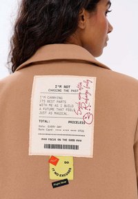 Žena v béžovém kabátu s motivujícími látkovými nášivkami na zádech, obsahujícími pozitivní citáty a dekorativní textové prvky.