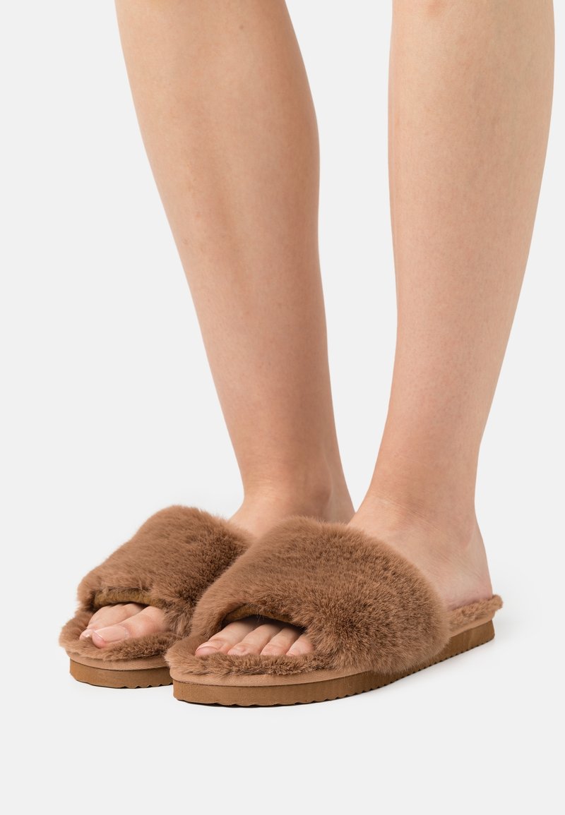 Pieds portant des chaussons marron ouverts à l'avant, avec des brides épaisses en fourrure et des semelles matelassées assorties, sur fond blanc.