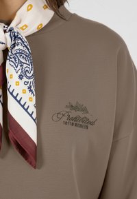 Brun bomullstopp med ribbet halslinning, kombinert med et mønstret silkesjal med marineblå, gul og burgunder aksenter. Bestikket logo detaljer.