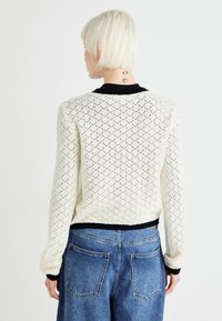 Pulover crem din tricot cu un model din dantelă, guler și mansete negre cu un model în relief, purtat cu blugi din denim albaștri cu talie înaltă.