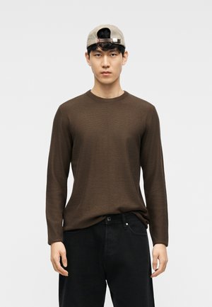 Jeune homme portant une chemise marron à manches longues, un pantalon noir et une casquette beige portée à l'envers, debout contre un fond clair uni.