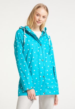 Junge Frau trägt eine hellblaue, gepunktete Regenjacke mit Kapuze und weißen Kordeln, steht vor einem schlichten weißen Hintergrund.