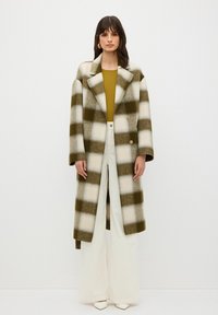 Cappotto lungo a quadri verdi e bianchi con rever oversize, due bottoni e una finitura morbida e testurizzata. Abbinato a un top senape e pantaloni bianchi.