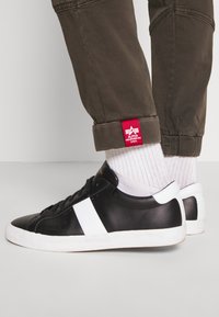 Zwarte leren sneakers met een witte zijstreep. Draag ze met geribbelde witte sokken en olijfgroene broekspijpen met een opgerolde rand en een rood etiket.