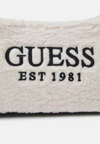 Crèmekleurige, getextureerde stof met een zwart geborduurd "GUESS"-logo en de tekst "EST 1981", met een zachte, pluizige uitstraling.