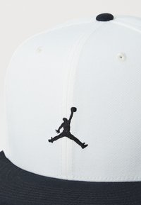 Casquette de baseball blanche avec une visière noire, ornée d'un logo Jumpman noir brodé sur le devant. Design structuré avec des trous de ventilation.