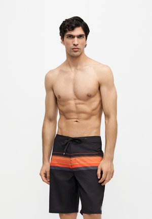 Skjorteløs mand med mørkt hår står iført sorte badeshorts med et orange og gråt stribet bånd over hofterne.