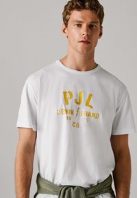T-shirt blanc en coton avec une impression jaune audacieuse indiquant "PJL DENIM BRAND CO. 1973", doté d'un col ras du cou classique et d'une coupe décontractée. Une ceinture verte à nouer en accent.