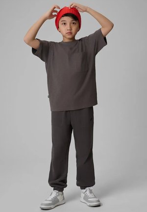 Młoda osoba poprawia czerwoną czapkę, ubrana w szary oversize t-shirt, dopasowane spodnie i białe trampki, stoi na jednolitym szarym tle.