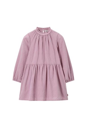 Enfant aux cheveux blonds bouclés tournant dans une robe rose avec un col rouge à motifs, des chaussettes violettes et des chaussures à boucle beige sur fond beige.
