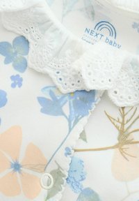 Biała tkanina z kwiatowym wzorem w delikatnym odcieniu niebieskiego i brzoskwiniowym, z koronkowym kołnierzykiem i małym guzikiem. Widoczna metka "NEXT baby".