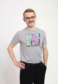 Šedé bavlněné tričko s logo v barevném stylu tie-dye, na kterém je nápis "MTV" a "MUSIC TELEVISION". Nosit se s černými kalhotami a opaskem.