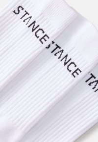 Meias brancas com nervuras, com o texto "STANCE" em preto impresso verticalmente, apresentando um punho sem costura e uma textura suave por todo o comprimento.