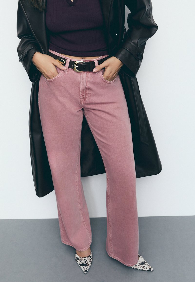 Roze široke traperice s prednjim džepovima, visokog struka, prošivanim detaljima, uparene s crnim remenom i šiljastim cipelama s uzorkom.