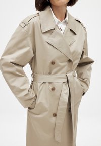 Beige trench coat valmistatud puuvilla segufabrist, millel on kahekordne eest, suured kraed ja vöökoht seotav rihm.
