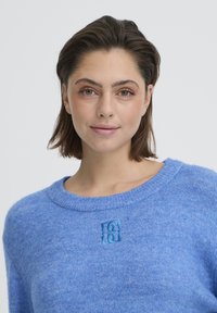 Pull bleu en tricot avec un col rond, présentant un détail de logo texturé dans une teinte de bleu plus claire à l'avant. Texture douce et légèrement duveteuse.