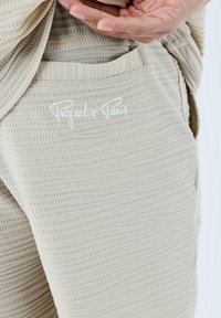 Joggers de tejido texturizado beige con un patrón en relieve, que cuentan con un bolsillo lateral y el logo bordado "Project x Paris" en blanco.