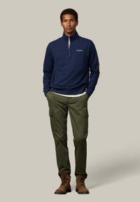 Sudadera con cremallera en el cuello de color marino con logo, combinada con pantalones cargo oliva y botas marrones. Tejido texturizado, corte relajado y bolsillos funcionales.