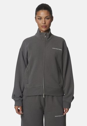 Donna che indossa una giacca con zip grigio scuro e pantaloni abbinati con logo "GOODFORNOTHING" sul petto e sulla coscia, in piedi su sfondo bianco.
