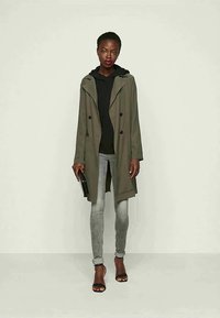 Trench coat verde-azeitona com um corte descontraído, design de duplo peito, colarinho largo, combinado com um hoodie preto e jeans skinny cinzentos.