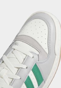 Detailní záběr na bílé tenisky s šedými semišovými a koženými prvky, zelenými pruhy, perforovanou špičkou a bílými tkaničkami.