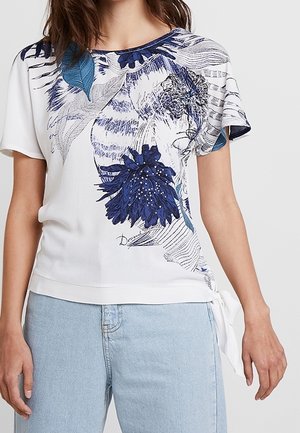 Frau trägt ein weißes Kurzarmoberteil mit blauem Blumen- und Tigerprint, kombiniert mit hellblauen Jeans, stehend vor einfachem Hintergrund.