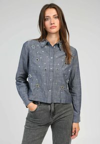 Chemise en denim, bleu clair, au design court. Présente des embellissements floraux et des accents métalliques argentés, avec une fermeture à boutons et des manches longues.