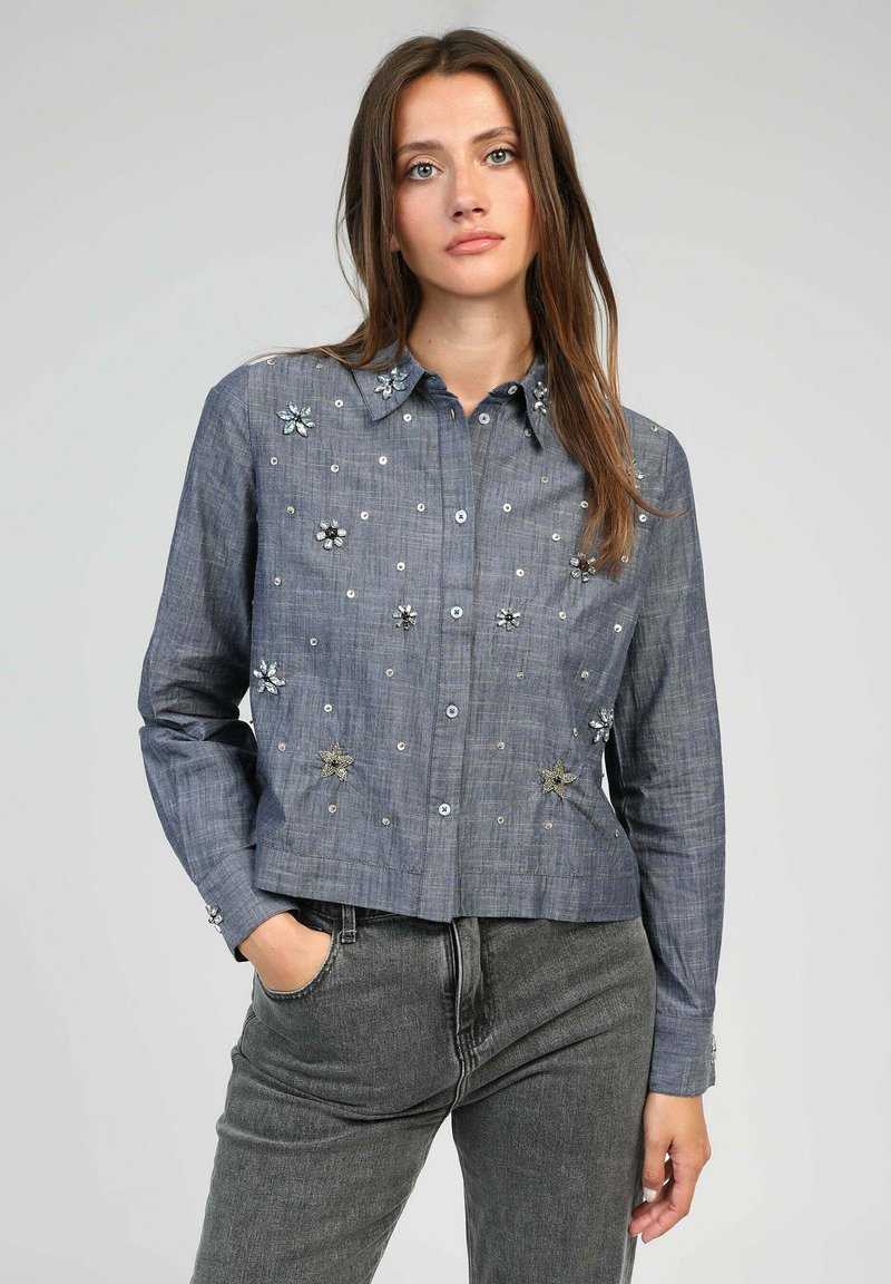 Chemise en denim, bleu clair, au design court. Présente des embellissements floraux et des accents métalliques argentés, avec une fermeture à boutons et des manches longues.