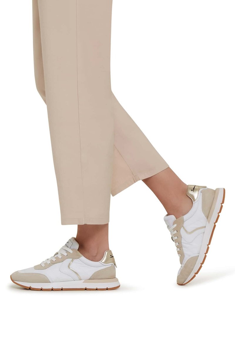 White and beige sneakers with suede accents and gold hardware. Lace-up design with a textured sole. Worn with cropped beige pants.