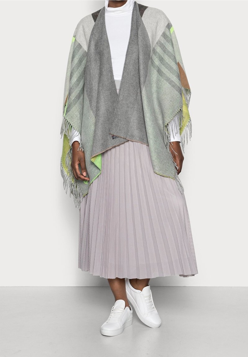 Personne portant une jupe plissée gris clair, un col roulé blanc, des baskets blanches, et un châle à franges gris et vert drapé sur les épaules.