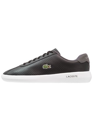 Baskets noires perforées avec semelle blanche, logo crocodile vert sur le côté et texte "LACOSTE" sur la semelle et la languette du talon.