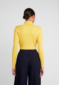 Maglione giallo a costine con collo alto e maniche lunghe, abbinato a pantaloni a vita alta di un blu navy scuro con righe verticali.