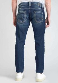 Jeans de mezclilla azul oscuro con un corte ajustado, que presentan detalles en los bolsillos traseros, un ligero desgaste y una cintura estándar. Llevados con zapatillas blancas.