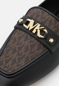 Gros plan sur un mocassin en cuir noir avec tissu marron à motifs et logo doré Michael Kors MK sur le dessus à l’avant.