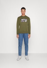 Sudadera verde oliva con diseño gráfico "JACK & JONES" en el frente, combinada con jeans de mezclilla azules y zapatillas blancas. Conjunto sencillo y casual.