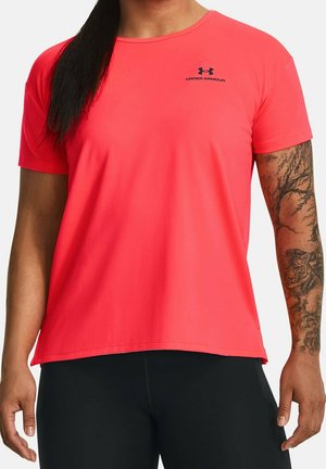 Person iført en lys pink Under Armour t-shirt og sorte bukser med tatoveret højre arm; baggrunden er ensfarvet og lys.