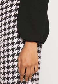 Main reposant sur un tissu à motif pied-de-poule noir et blanc, portant une longue manche bouffante noire avec un poignet élastique.