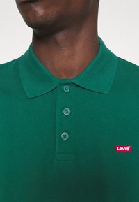 Homme portant un polo vert à trois boutons avec un petit logo rouge Levi's sur la poitrine, faisant face à l'avant.