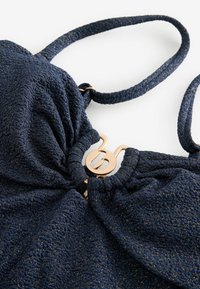 Top de bikini azul marino texturizado con tirantes ajustables y un centro fruncido. Presenta un detalle de cierre en tono dorado para abrochar.